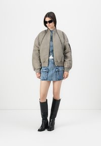 Donna con giacca bomber grigia oversize, mini gonna di jeans con tasche, stivali neri sopra il ginocchio e occhiali da sole scuri, su sfondo bianco.