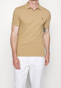 Hnědá bavlněná polo košile s klasickým límcem, krátkými rukávy a malým logem na hrudi. Spárována s bílými kalhotami.
