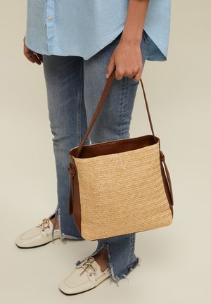 Person trägt hellblaues Hemd und Jeans mit ausgefranstem Saum und hält eine gewebte beigefarbene Tragetasche mit braunen Lederstreifen und Besatz.