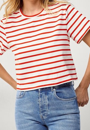 Kobieta w białym krótkim t-shircie z poziomymi czerwonymi paskami i wysokim stanem niebieskich dżinsów, ręce spoczywają na biodrach.