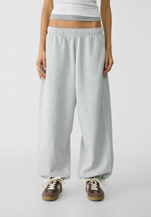 Pantaloni da tuta grigi con cintura elastica, design a gamba larga e cuciture laterali bianche, abbinati a sneaker marroni con lacci chiari.