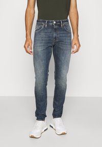 Blaue Jeans im Skinny-Fit mit ausgewaschenen Kniedetails, klassischem Fünf-Taschen-Design und Metallelementen, kombiniert mit weißen Sneakers.