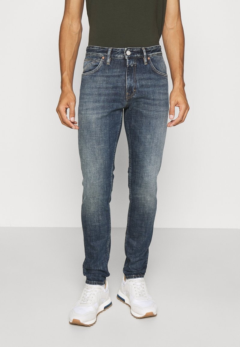 Blaue Jeans im Skinny-Fit mit ausgewaschenen Kniedetails, klassischem Fünf-Taschen-Design und Metallelementen, kombiniert mit weißen Sneakers.