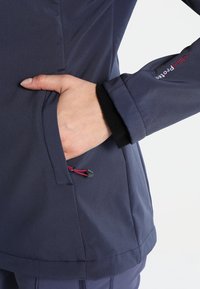 Hand in the pocket of a navy blue jacket with fleece lining detail and "Clime Protect" embroidered on the sleeve cuff.
