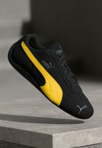 Sneaker atletica nera e gialla con una morbida texture in suede, caratterizzata da un design elegante, logo Puma e pannelli laterali testurizzati per una migliore aderenza.