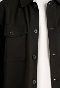 Zwart overhemd met knopen, een puntige kraag, één borstzak en zwarte knopen; gemaakt van een gestructureerde stof, getoond tegen een witte achtergrond.