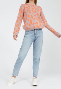 Strickpullover in Hellpink mit orangenem abstraktem Muster, kombiniert mit hellblauen schmal geschnittenen Jeans und weißen Sneakers.
