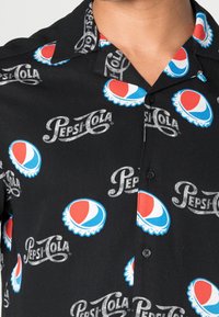 Short-sleeve black shirt features a repeated print of the Pepsi logo and bottle caps in red, blue, and white. Fabric has a smooth texture.