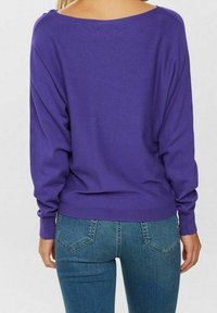 Lila Langarm-Pullover mit weitem Ausschnitt und gerippten Bündchen, kombiniert mit enger blauer Jeans mit Gesäßtaschen.