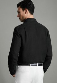Schwarzes Leinenhemd mit einem geknöpften Kragen, langen Ärmeln und einem lässigen Schnitt, kombiniert mit weißen Hosen und einem schwarzen Gürtel.
