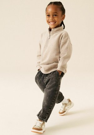 Child with braided hair smiling, wearing beige zip-up sweater, black jeans, and beige sneakers, walking with hands in pockets on light background.