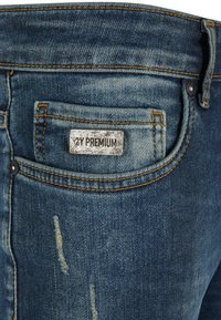 Tumed sinine denimist teksapüksid, millel on kulumise detailid, tagaküljel on sildiga tasku, millel on kirjas "2Y PREMIUM", ja kontrastse kollase õmblusega.