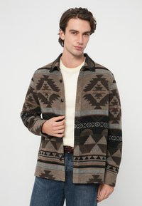 Camisa de franela de lana con un patrón geométrico en marrón, negro y gris, con cuello de solapa, cierre frontal con botones y un corte relajado.