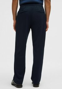 Persona vistiendo pantalones rectos de color azul marino oscuro y zapatos de vestir negros, de pie con las manos relajadas a los lados, vista posterior.
