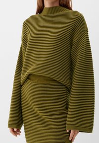 Pull en maille côtelée vert olive avec col haut, coupe courte et manches larges, assorti à une jupe côtelée assortie.