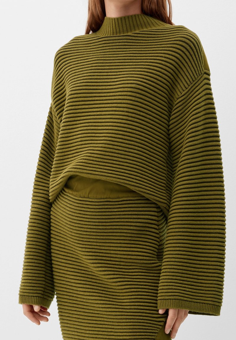 Pull en maille côtelée vert olive avec col haut, coupe courte et manches larges, assorti à une jupe côtelée assortie.