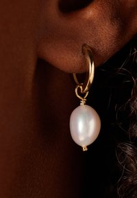 Gouden oorbel met een gladde, ovale, witte parel als hanger. Gestructureerde huid en donker haar zichtbaar. Nader in beeld, toont detail.