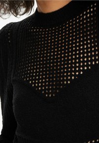 Czarny dzianinowy sweter z wysokim dekoltem i geometrycznym wzorem okrągłych wycięć w górnej części. Szczegóły w teksturowanym materiale.