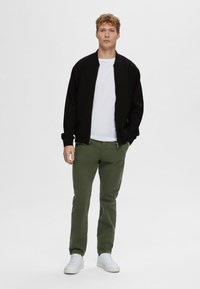 Zwarte bomberjack over een wit T-shirt, olijfgroene broek en witte sneakers. Glad materiaal met ribbelmanchetten en tailleband.