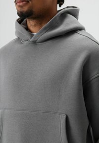 Grijze hoodie van zachte stof, met een grote voorkantzak, een ontspannen pasvorm en een capuchon met trekkoord. Zachte textuur met subtiele naden.