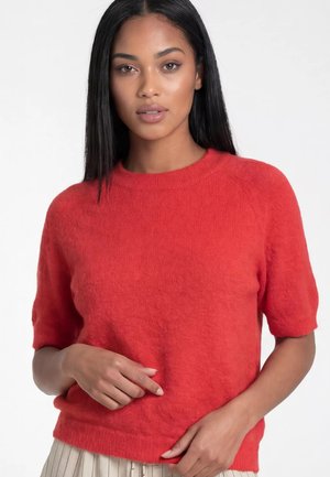 Kobieta z prostymi czarnymi włosami, ubrana w czerwony sweter z krótkim rękawem i jasną plisowaną spódnicę na jednolitym tle.