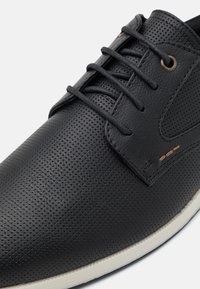 Zapato de cuero negro perforado con un diseño clásico de cordones, suela de color claro contrastante y discretos detalles de costura.