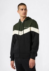 Uomo che indossa una felpa con zip nera e verde scuro, con una striscia a chevron bianca, in piedi contro uno sfondo chiaro uniforme, che guarda di lato.