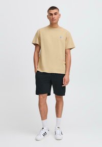 Camiseta de algodón beige con mangas cortas y un diseño bordado en el pecho, combinada con pantalones cortos negros y zapatillas deportivas blancas.