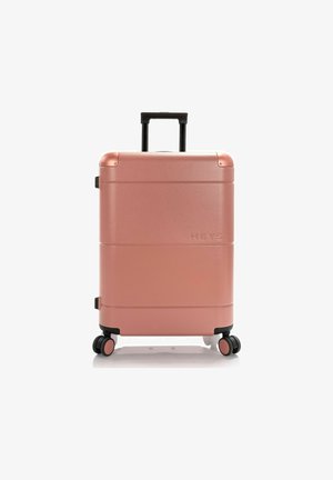Valigia rigida color oro rosa con finitura liscia, ruote nere e manico estraibile. Presenta il nome del brand "HEYS" impresso di lato.
