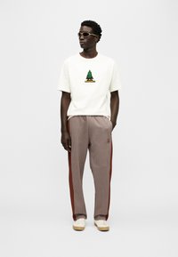 Λευκό κοντομάνικο μπλουζάκι με πράσινο γραφικό δέντρο· καρό καφέ παντελόνι jogging με λωρίδες στο πλάι· λευκά αθλητικά παπούτσια· στρογγυλά γυαλιά ηλίου.