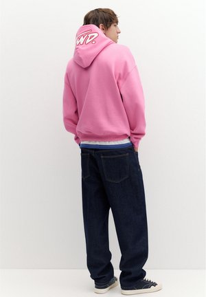 Giovane uomo che indossa una felpa rosa con cappuccio e scritta bianca sul cappuccio, jeans blu scuro e sneakers nere con suola bianca, in piedi contro un muro semplice.
