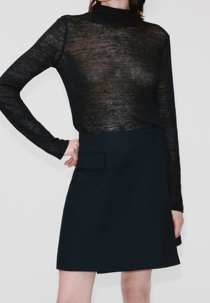 Mujer vistiendo una blusa negra transparente de manga larga metida en una falda negra de talle alto con un bolsillo con solapa en la parte delantera.