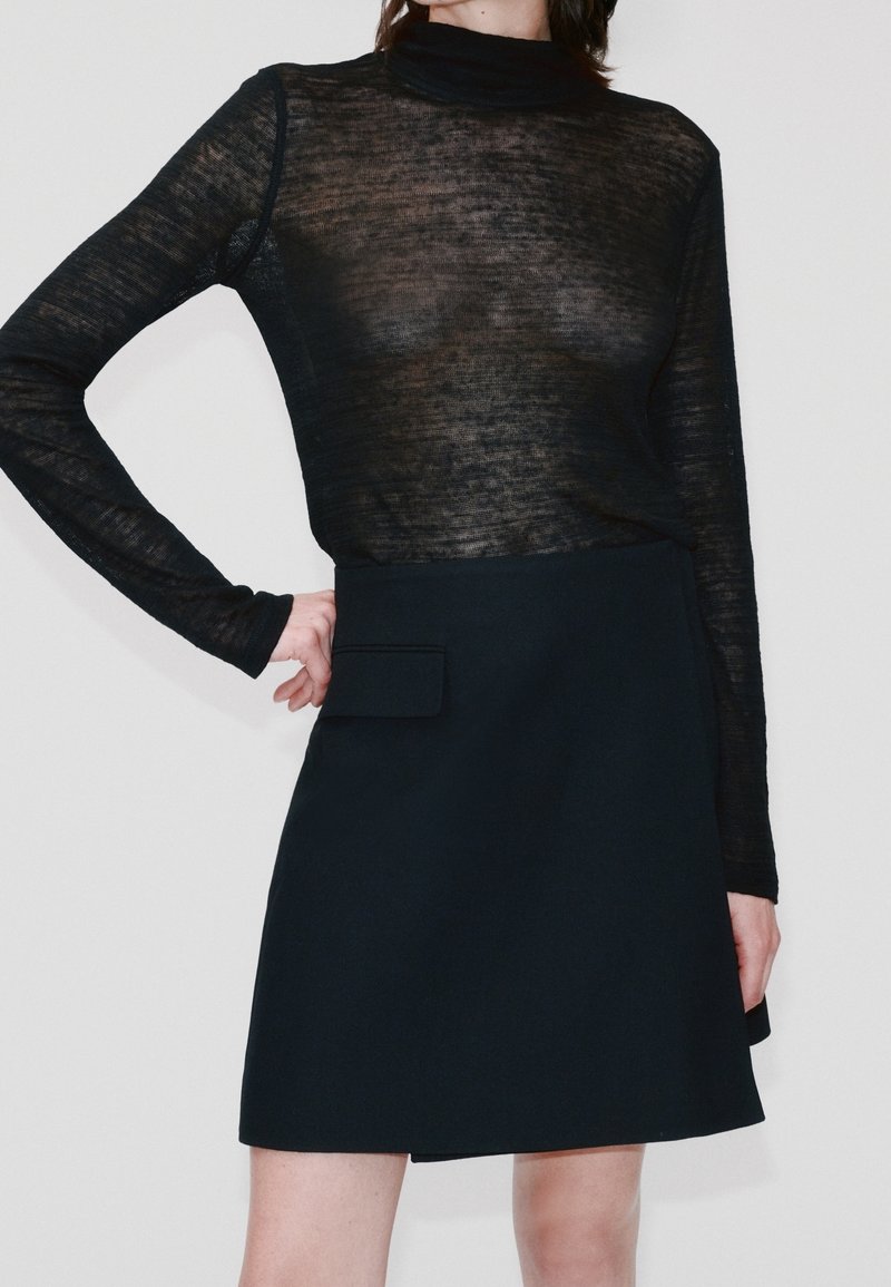 Femme portant un haut noir transparent à manches longues, rentré dans une jupe noire taille haute avec une poche à rabat à l'avant.