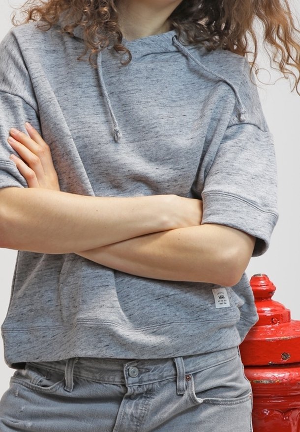 Sudadera corta de manga corta gris con una tela texturizada, capucha con cordón y una pequeña etiqueta de la marca en el dobladillo, combinada con jeans gris claro.