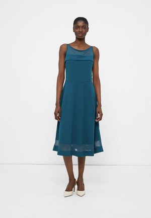 Γυναίκα που φοράει πράσινο μπλε αμάνικο midi φόρεμα με διαφανή λαιμόκοψη και τελείωμα, και λευκά μυτερά τακούνια, μπροστά σε απλό λευκό φόντο.