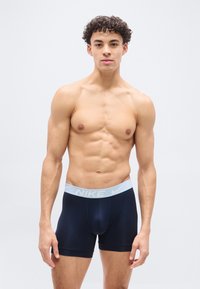 Άνδρας μοντέλο με σγουρά μαλλιά που φοράει μπλε-σκούρα boxer briefs της Nike με ανοιχτό μπλε ζωνάκι στη μέση, στέκεται μπροστά σε απλό φωτεινό φόντο.