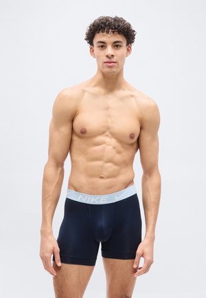 Άνδρας μοντέλο με σγουρά μαλλιά που φοράει μπλε-σκούρα boxer briefs της Nike με ανοιχτό μπλε ζωνάκι στη μέση, στέκεται μπροστά σε απλό φωτεινό φόντο.