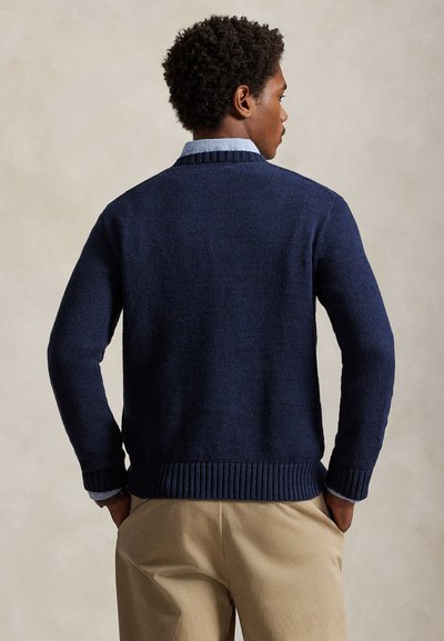 Man with curly hair wearing a navy blue sweater over a light blue collared shirt and beige pants, standing facing away with hands in pockets.