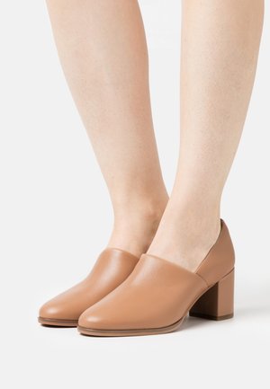 Chaussures à talon bloc beige avec une texture en cuir lisse et un design minimaliste, présentant un bout arrondi et une silhouette à côtés ouverts.