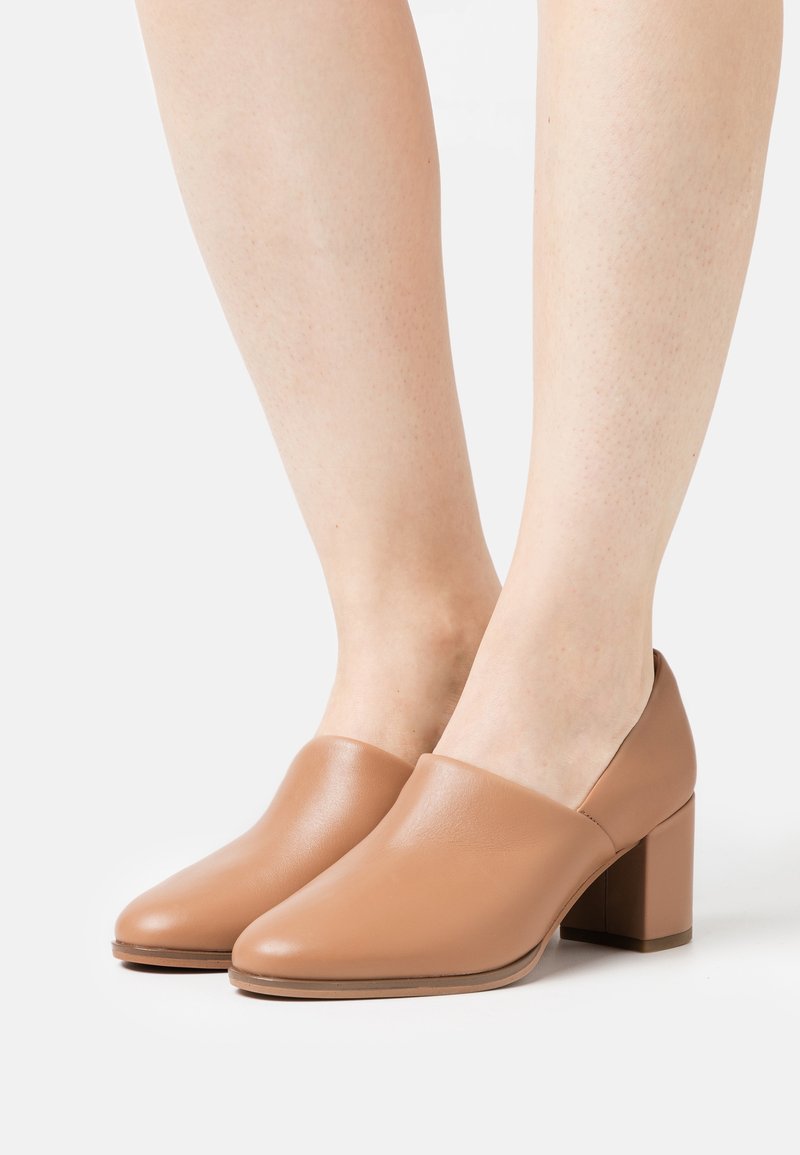 Chaussures à talon bloc beige avec une texture en cuir lisse et un design minimaliste, présentant un bout arrondi et une silhouette à côtés ouverts.