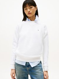 Sudadera blanca con puños y dobladillo acanalados, con un pequeño logo en el pecho, superpuesta sobre una camisa de color azul claro con cuello y combinada con jeans azules.