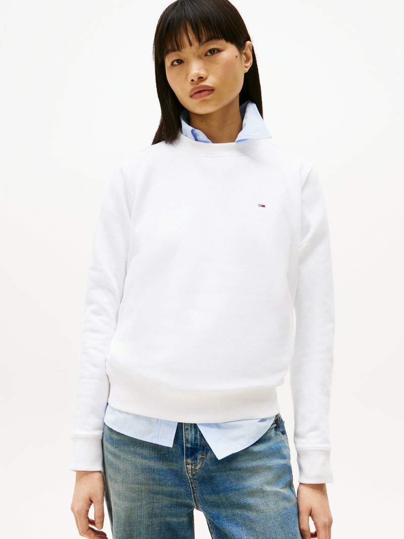 Sudadera blanca con puños y dobladillo acanalados, con un pequeño logo en el pecho, superpuesta sobre una camisa de color azul claro con cuello y combinada con jeans azules.