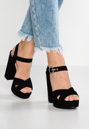 Czarne sandały na wysokim obcasie z krzyżowym paskiem, wykonane z zamszu, z okrągłą klamrą na pasku kostki i otwartym noskiem.