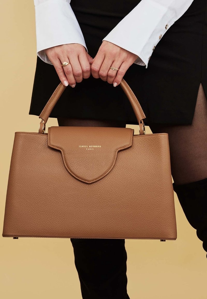 Personne portant une chemise blanche et une jupe noire tient un sac à main en cuir marron avec des poignées supérieures et des lettres dorées de la marque.