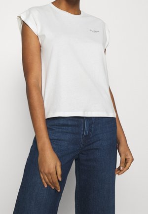 Persona vestida con una camiseta blanca sin mangas con un pequeño logo en el pecho y vaqueros azules de talle alto, de pie con los brazos relajados.