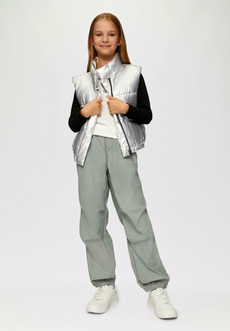 Zilveren puffervest met hoge kraag, zwarte shirt eronder en lichtgroene baggy broek. Witte sneakers en een ontspannen pasvorm in het algemeen.