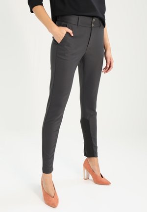 Femme portant un pantalon gris foncé ajusté avec des patchs noirs aux chevilles et des chaussures à talons hauts corail, une main dans la poche, debout sur un fond blanc.