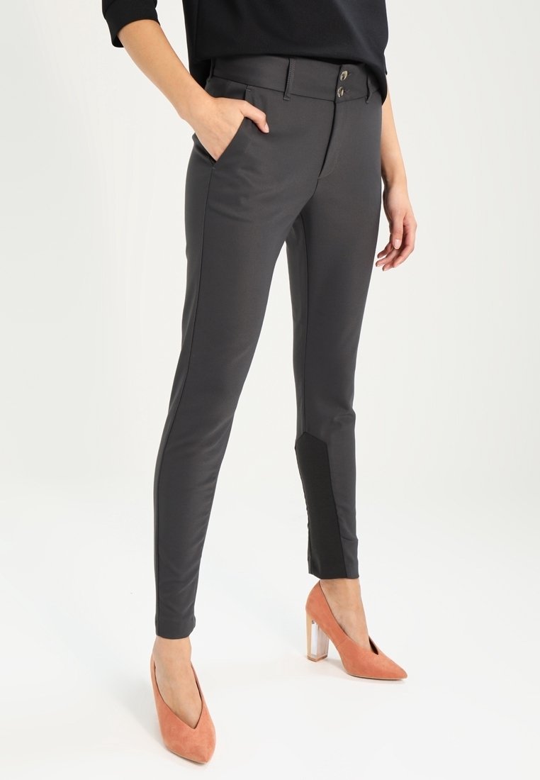 Femme portant un pantalon gris foncé ajusté avec des patchs noirs aux chevilles et des chaussures à talons hauts corail, une main dans la poche, debout sur un fond blanc.
