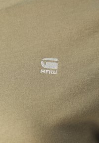 Olivengrønt teksturert stoff med en hvit sydd logo med ordet "RAW" under. Enkel, moderne design uten ekstra mønstre.