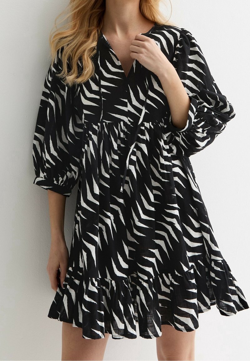 Femme portant une robe imprimé zèbre noir et blanc avec un col en V, des manches bouffantes et un ourlet volanté sur un fond uni.